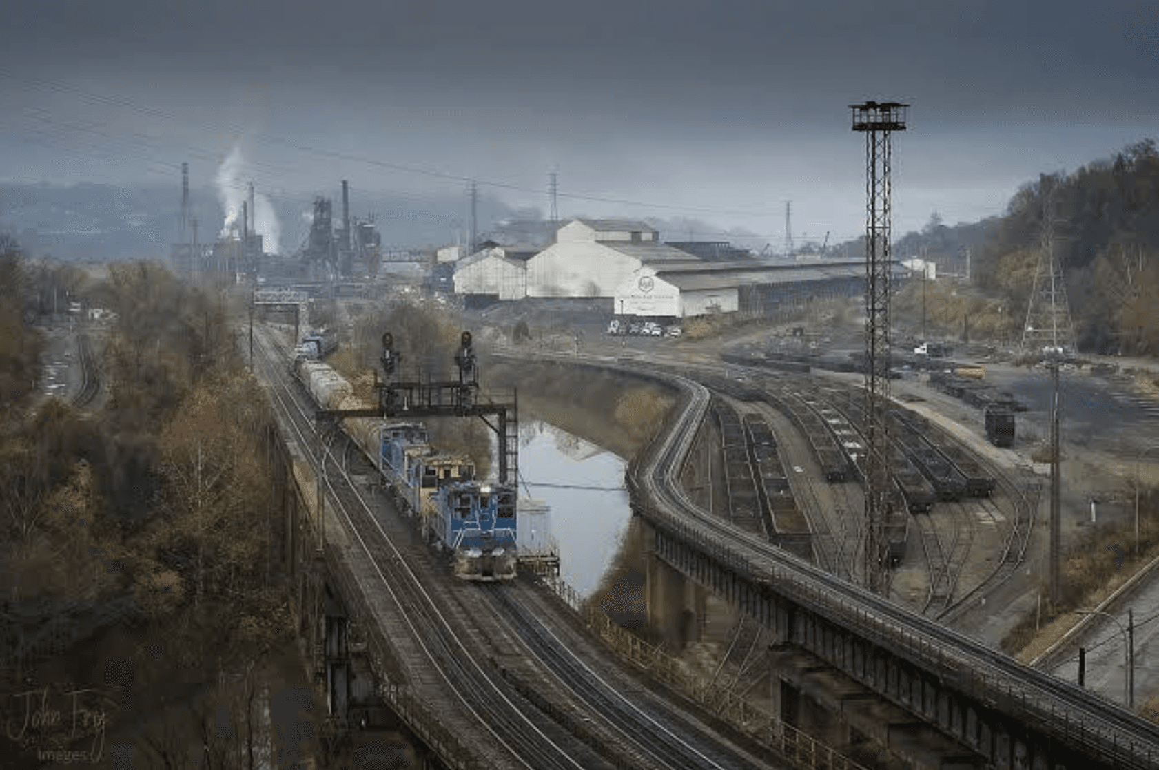 The image depicts an industrial railway yard with multiple tracks, a blue loading chute, smokestacks, and a landscape with trees.