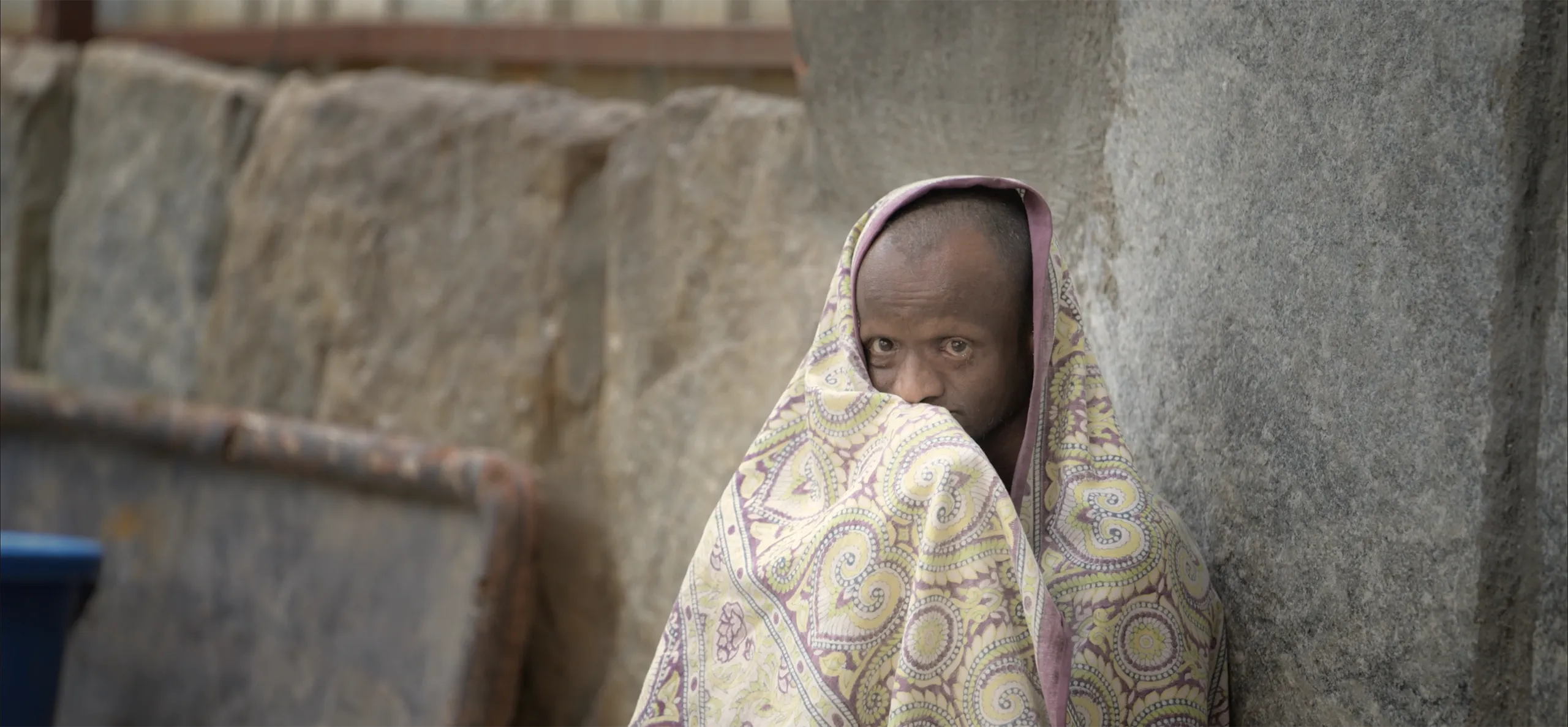 A poignant and intimate portrait of a person wrapped in a patterned blanket looking shyly at the camera.