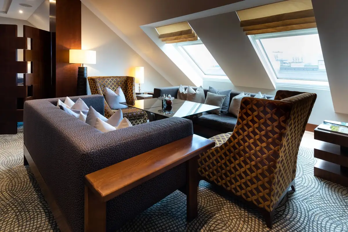 A cozy lounge with patterned armchairs and a gray sofa in a room with large slanted windows overlooking water.