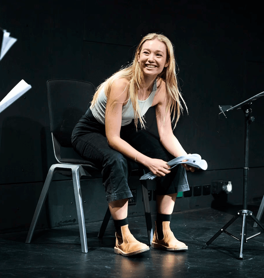 A young woman sits on a chair in a rehearsal studio, smiling as she holds a script. She leans forward with relaxed posture, surrounded by other performers holding pages, with a music stand and personal items on the floor nearby.