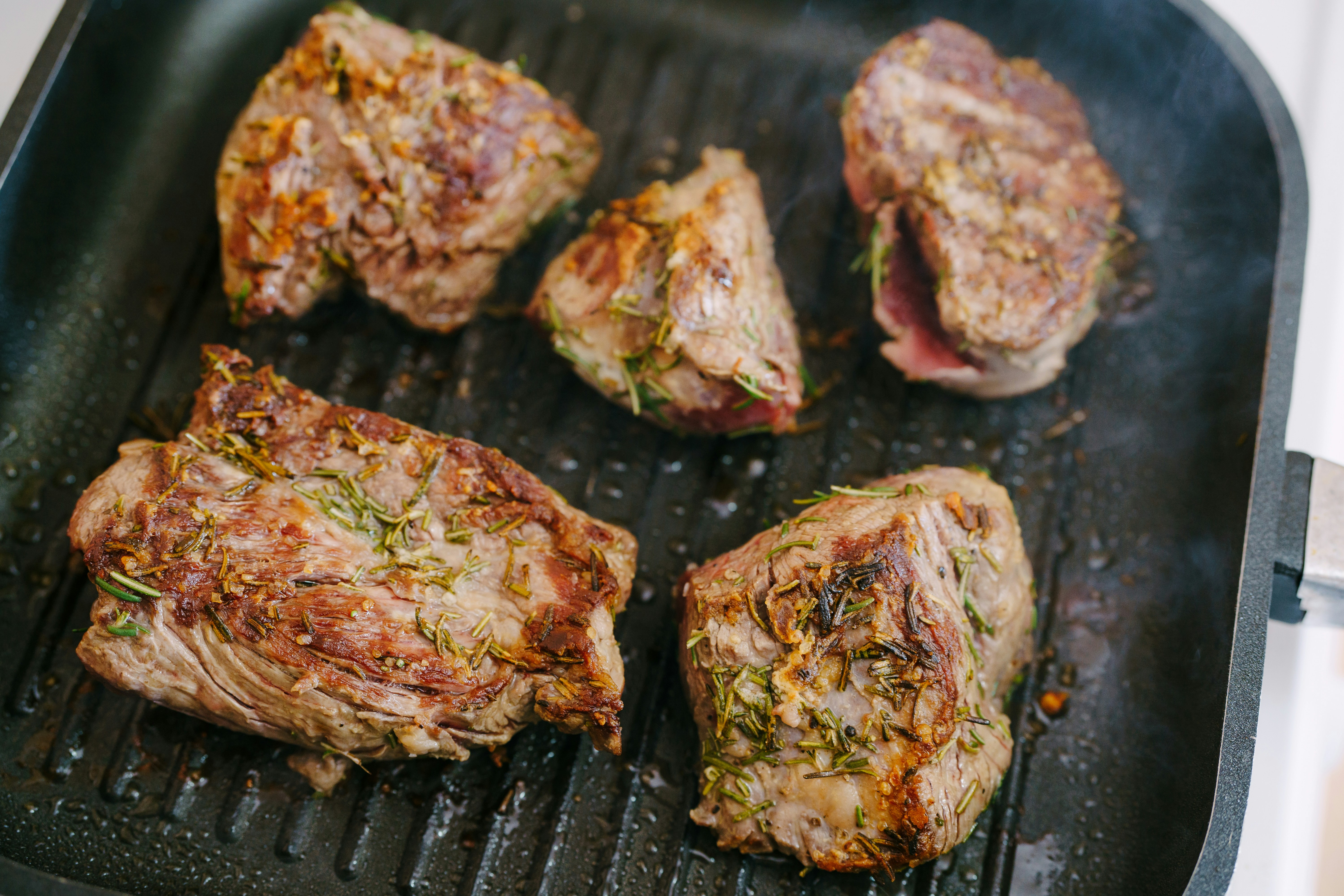 Five pieces of seasoned meat cooking on a grill pan.