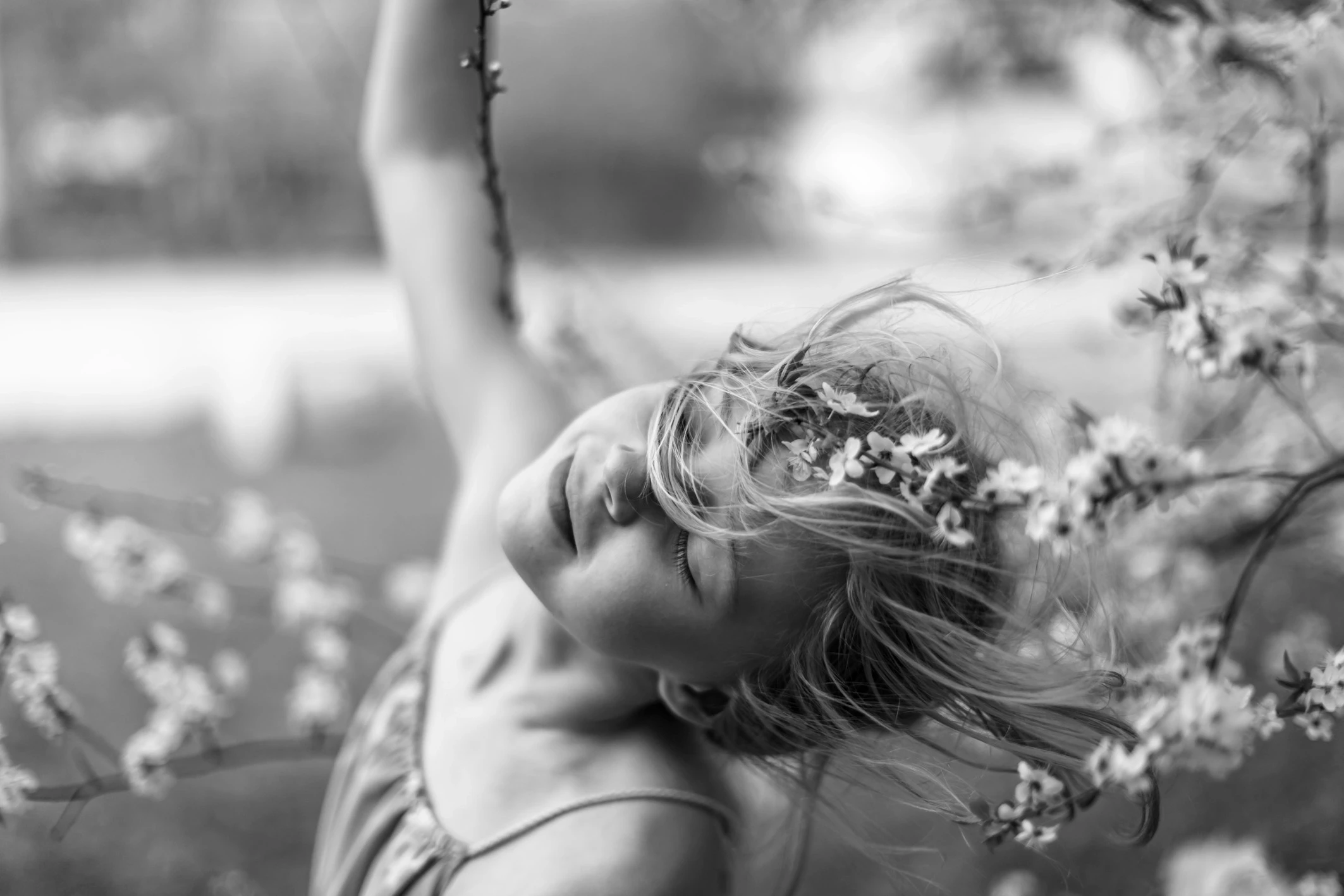 Czarno-białe zdjęcie młodej uśmiechniętej osoby z zamkniętymi oczami, głową odchyloną do tyłu i zmierzwionymi włosami wśród kwitnących gałęzi, tworząc radosny i pogodny nastrój.