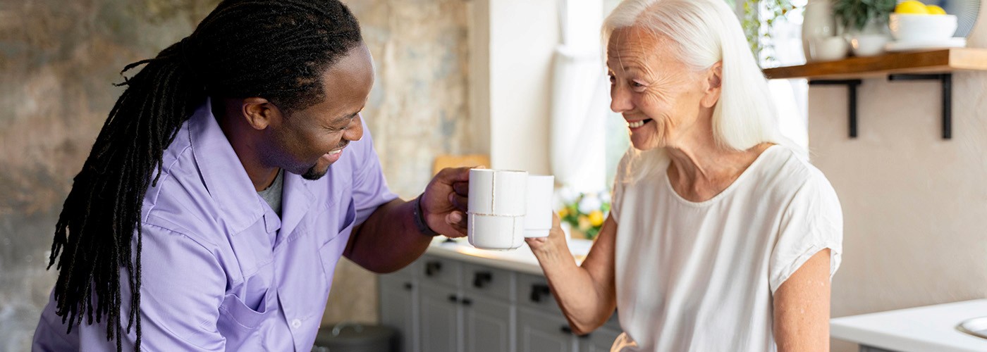 A male caregiver in a purple uniform and an elderly woman smiling while sharing a toast with coffee mugs in a kitchen