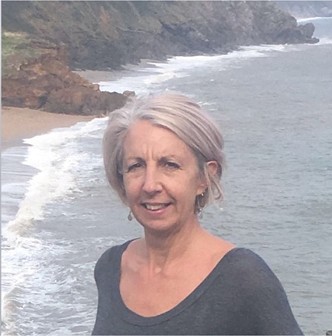 A smiling woman with short gray hair stands by the beach, with a coastline in the background.