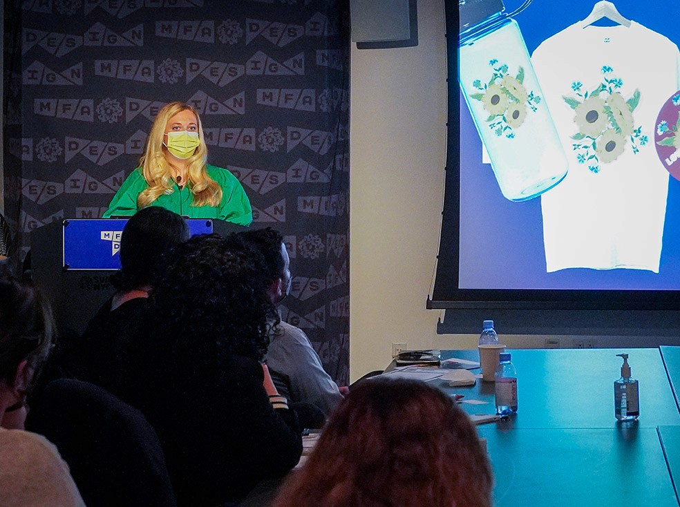A woman dressed in green is giving a speech and on the right side is an image projected with a t-shirt on a hanger, a jar, and other objects with flower graphics on them.