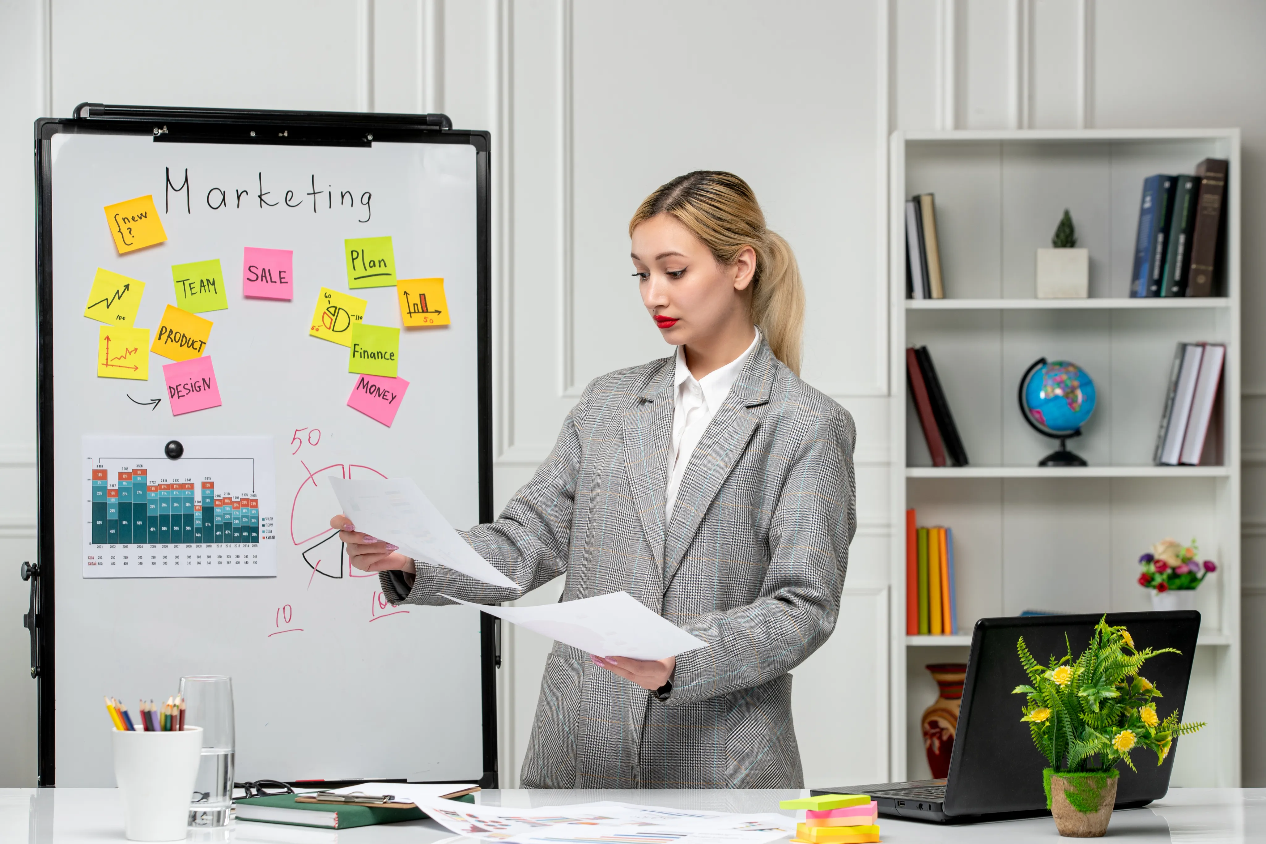 Person in a blazer presenting marketing plans with sticky notes and charts on a whiteboard in a modern office setting.