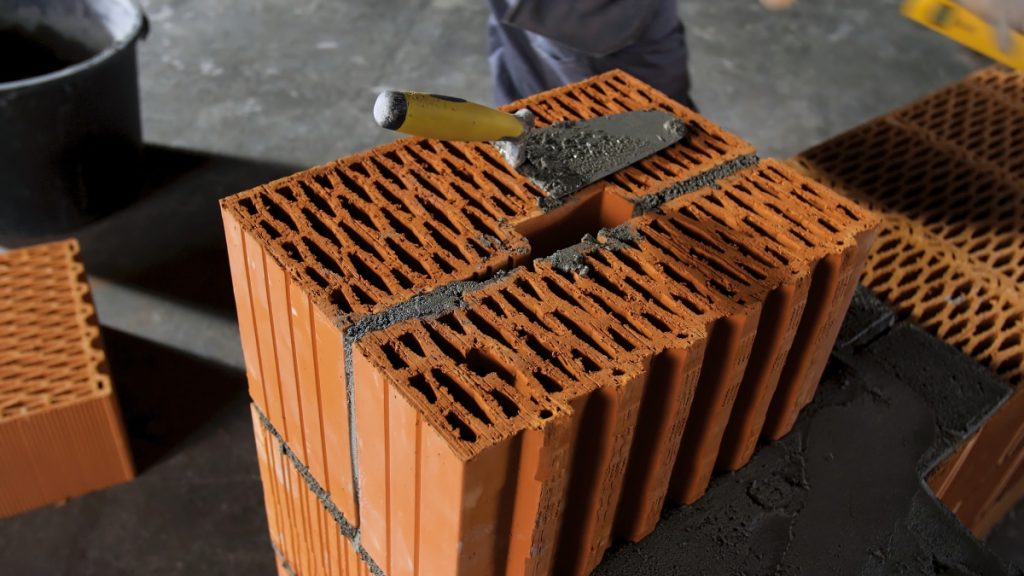 Bricklayer worker putting away extra cement with trowel putty knife on grey floor background. Stock footage. Man in gloves building a red brick wall.