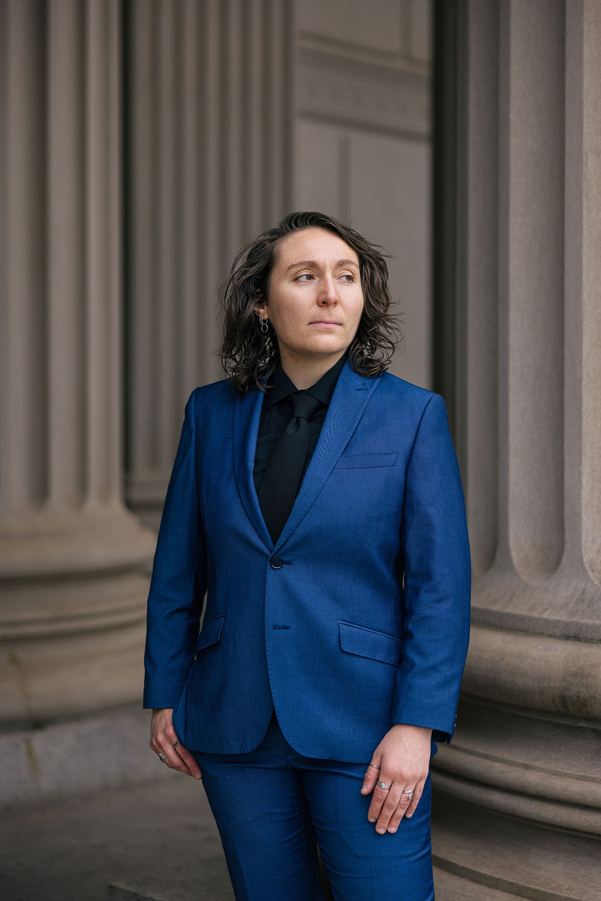 Omny Miranda Martone stands between large stone columns in a bright blue suit, looking off to the side.