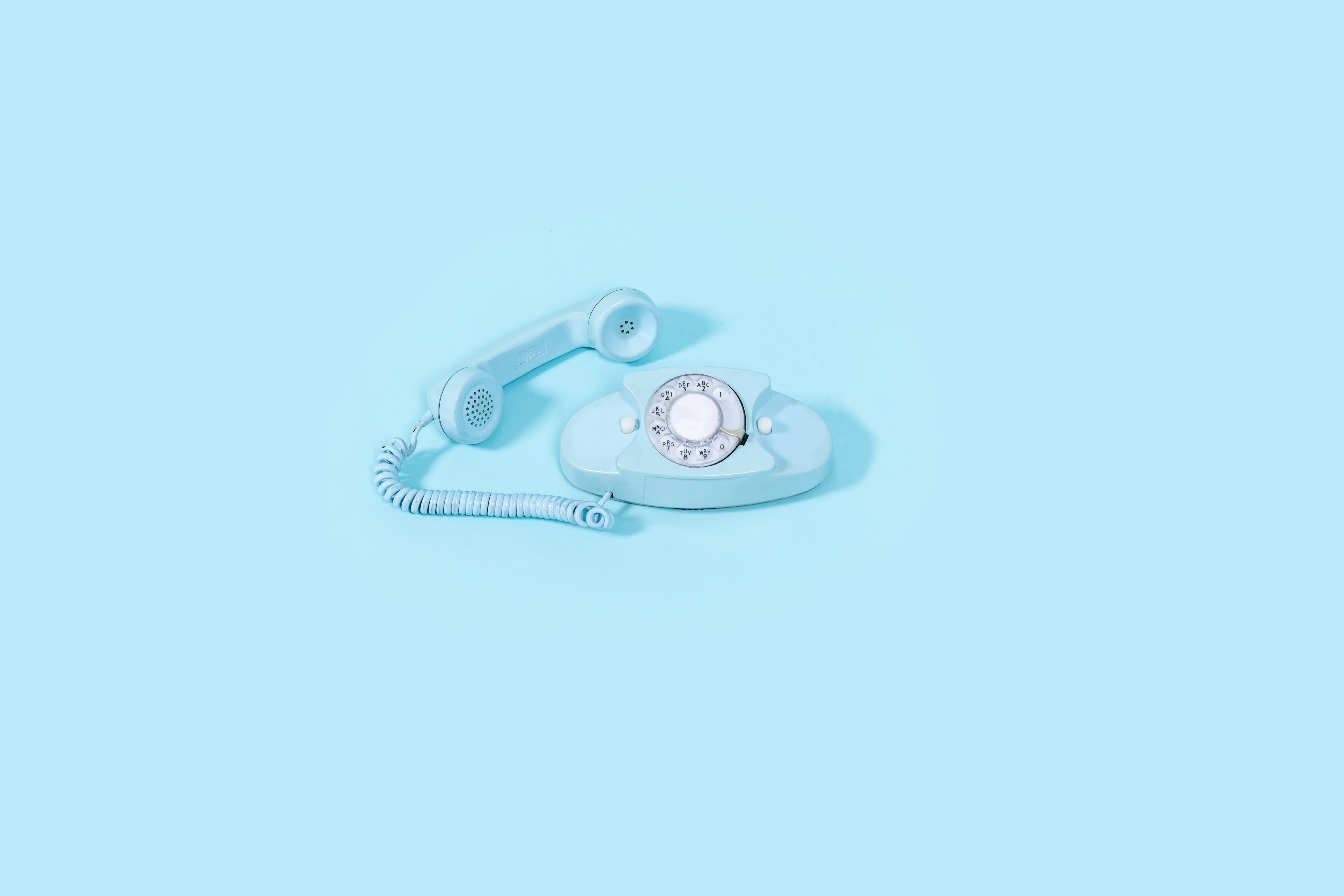 Telefon obrotowy ze słuchawką przymocowaną za pomocą zwijanego sznura. Zarówno telefon, jak i tło są w jaskrawym, błękitnym kolorze. - ilustracja dla artkułu na temat Chatbot rekrutacyjny: jak zautomatyzować komunikację z kandydatem