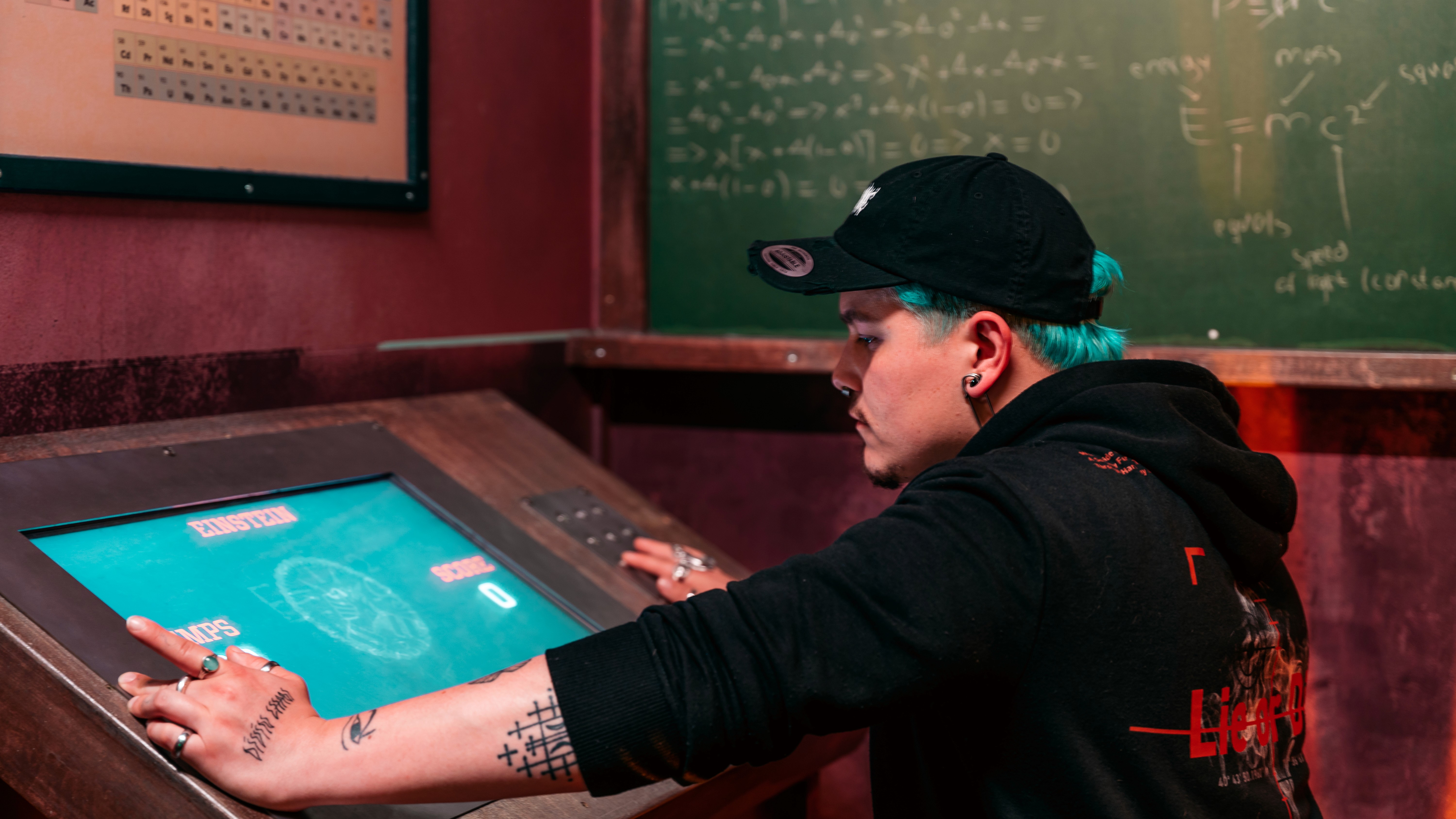 A young person with curly hair smiles at the camera while sitting in front of a laptop against a dark background.