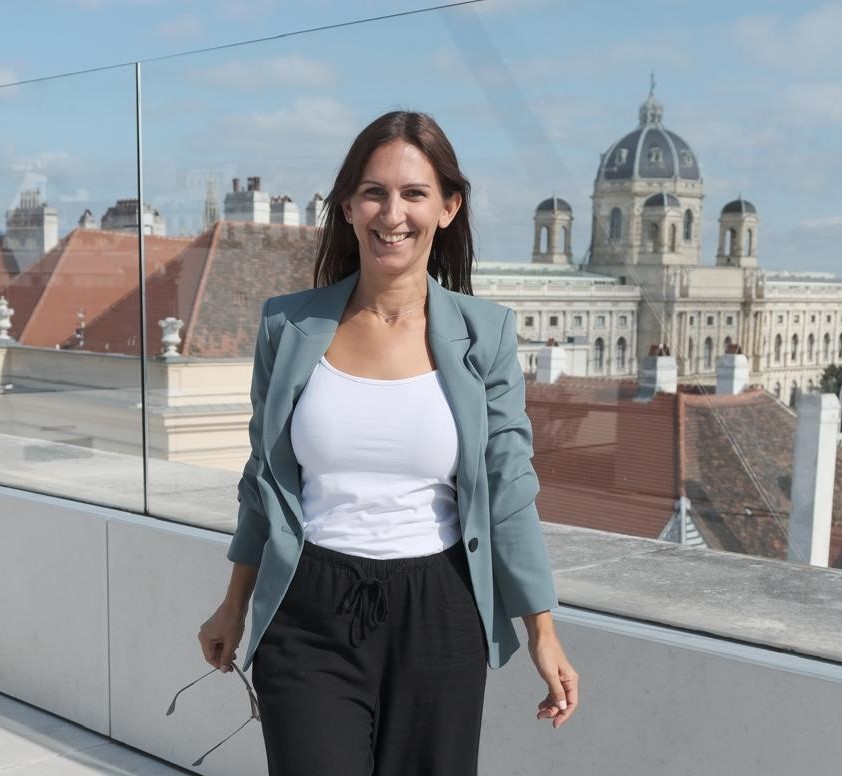 Eine lächelnde Frau mit langen Haaren steht im Freien vor einer städtischen Kulisse mit historischen Gebäuden; sie trägt ein helles Oberteil und eine dunkle Jacke.