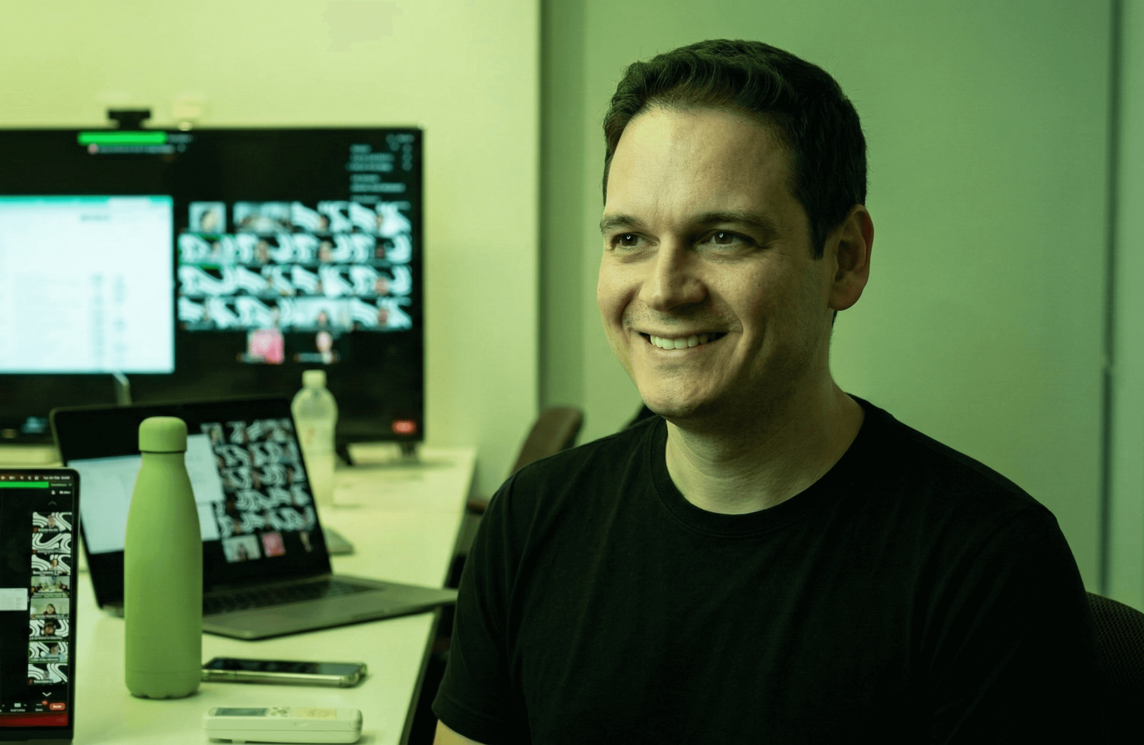 Pedro Pazitto smiling at a desk in a modern office, with a laptop and a large monitor displaying a video conference in the background.