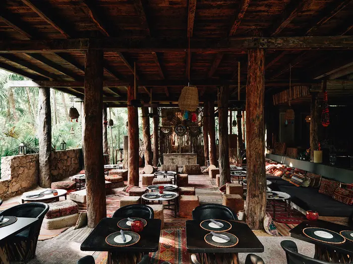 Mesas y sillas en lujosos tonos negros con lámparas colgantes y soportes de troncos de árboles en el restaurante mediterráneo llamado Macondo, rodeado de naturaleza y selva en Nômade Tulum, México.