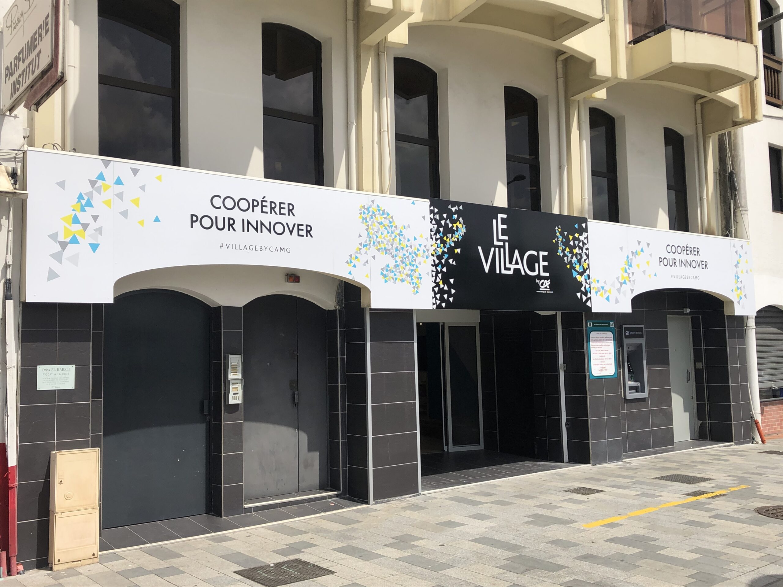 Vitrine avec l'enseigne « Le Village », flanquée de phrases motivantes et de motifs triangulaires colorés. Le décor dégage une atmosphère moderne et collaborative.