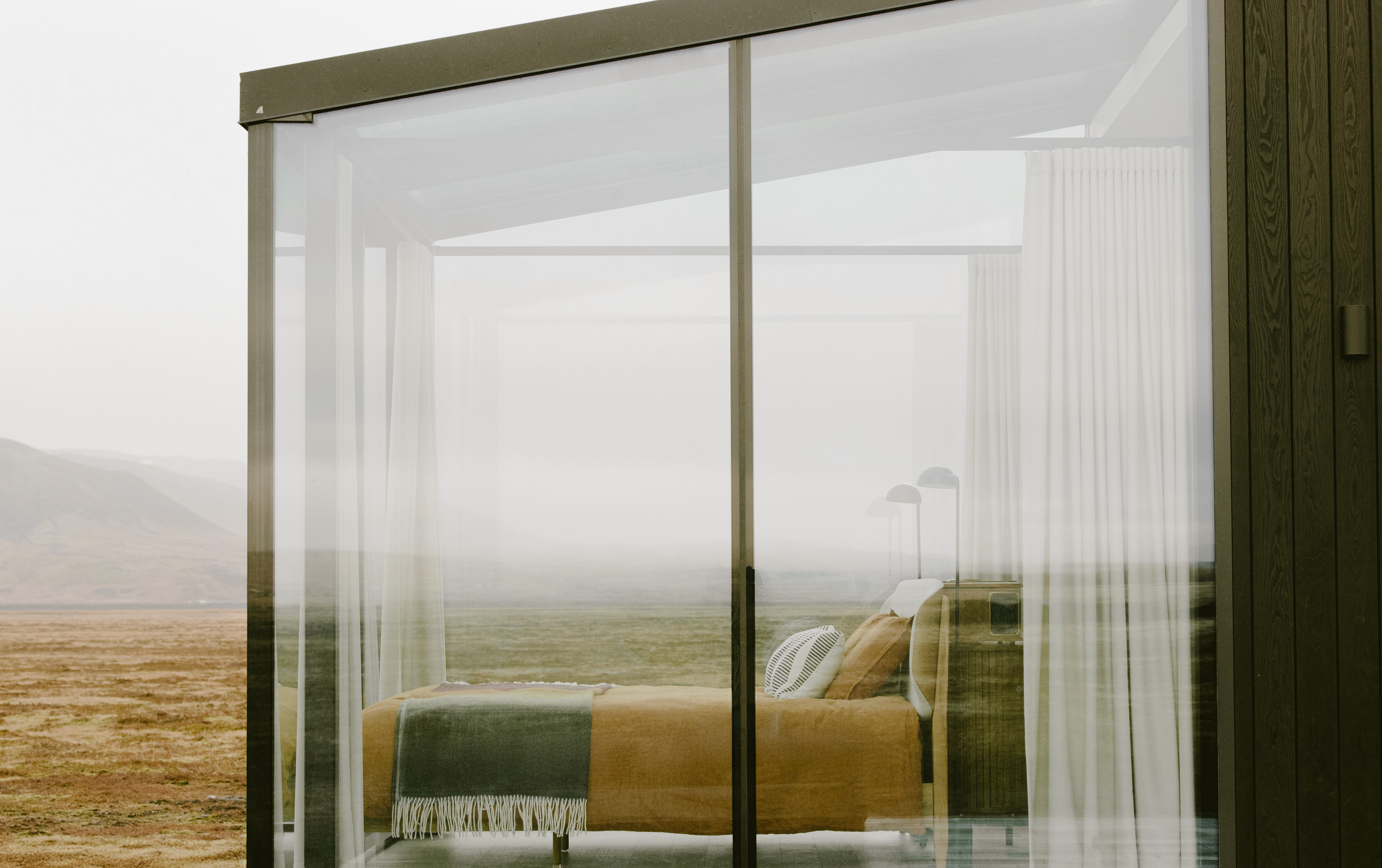 Exterior view of a modern cabin with floor-to-ceiling glass, revealing a minimalist bedroom with a low bed and soft textiles, set against an open, barren Icelandic landscape under a muted sky.