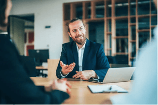 A woman sitting at a desk in a bright office, working on a laptop with notebooks, a cup, and a desk lamp beside her.