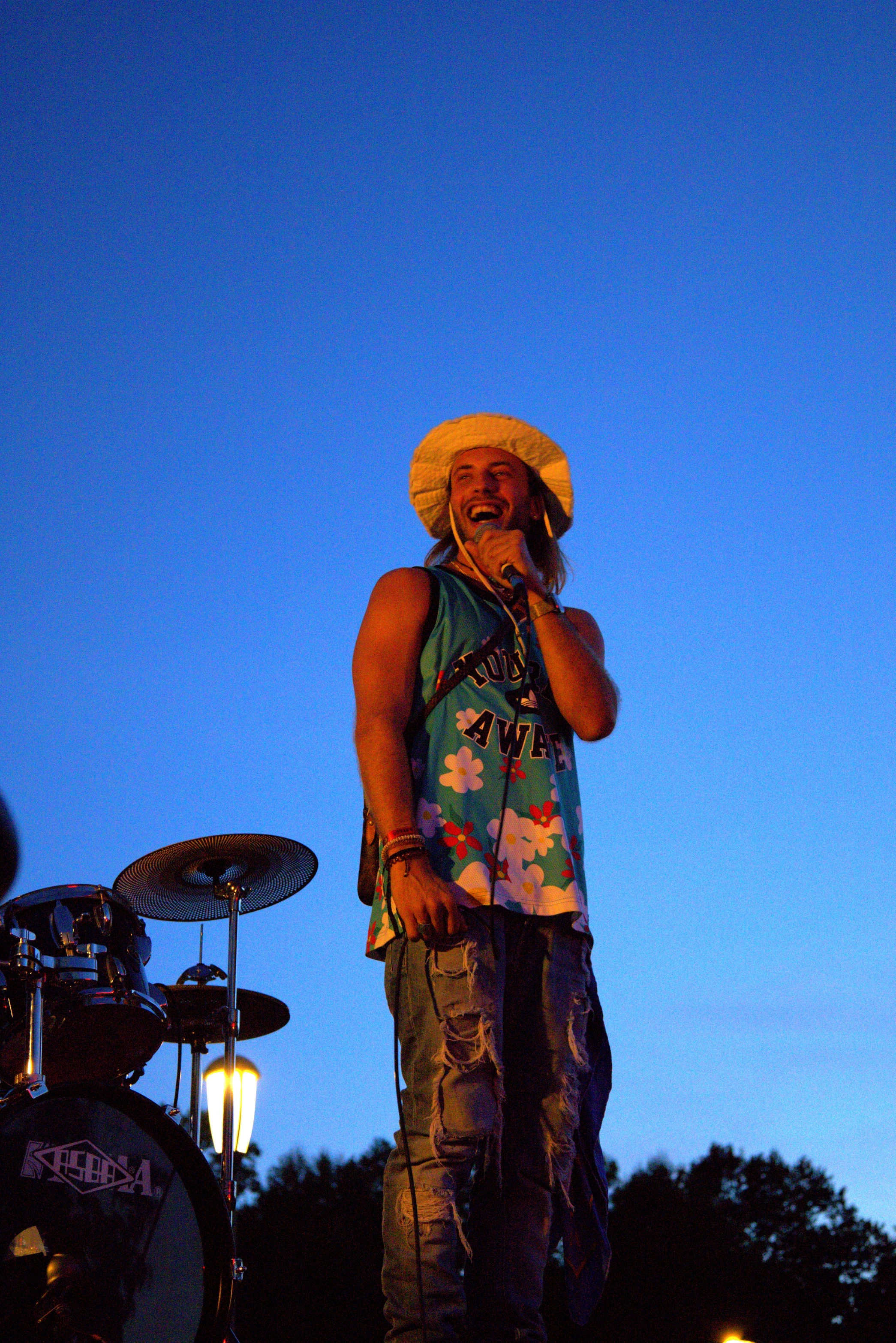 A performer stands on stage against a clear blue sky, dressed in vibrant colors and holding a microphone.