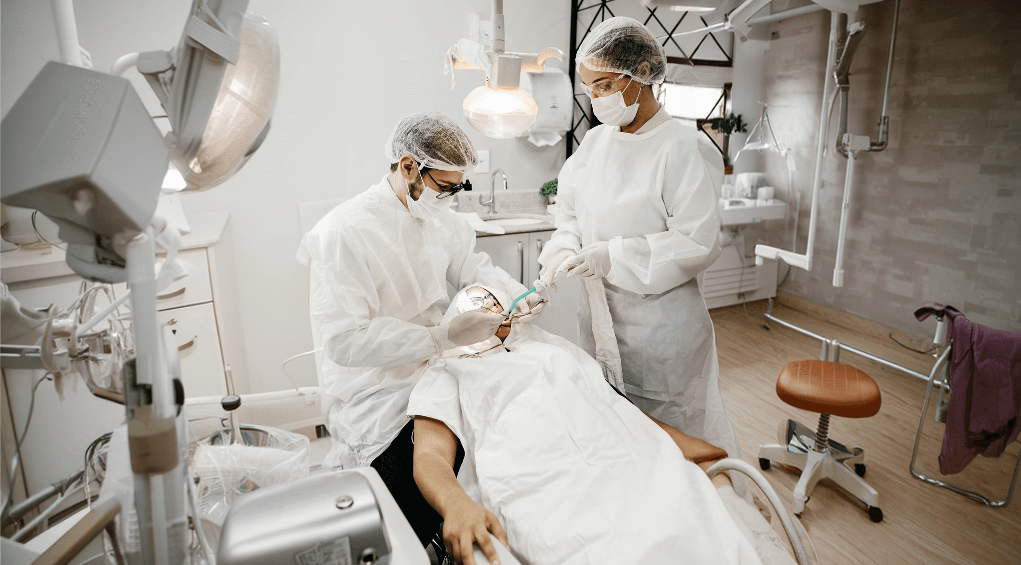 Two masked dental professionals in white uniforms performing a procedure on a patient under a bright examination light