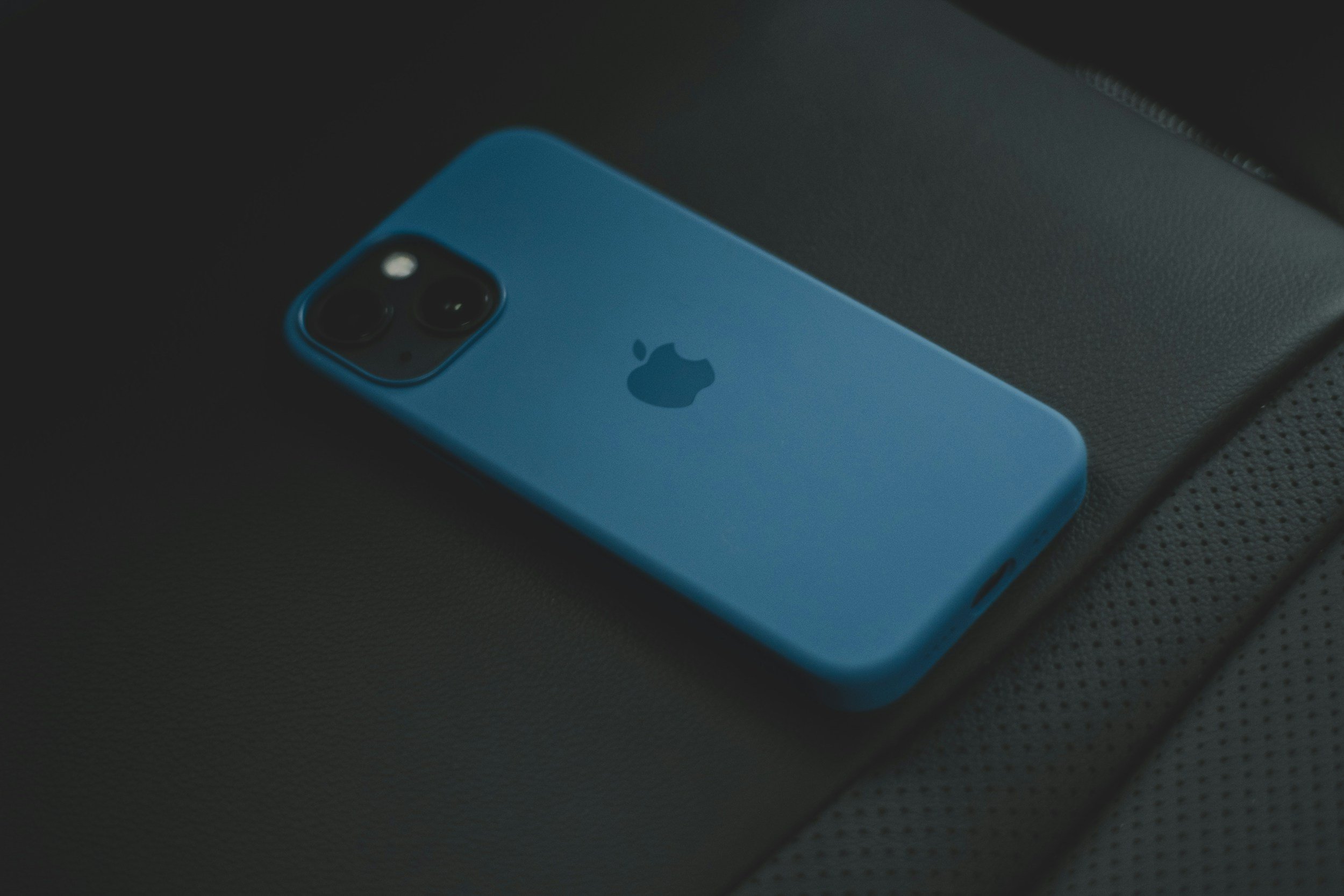 Blue iPhone with dual rear cameras, featuring the Apple logo, resting on a dark textured surface.