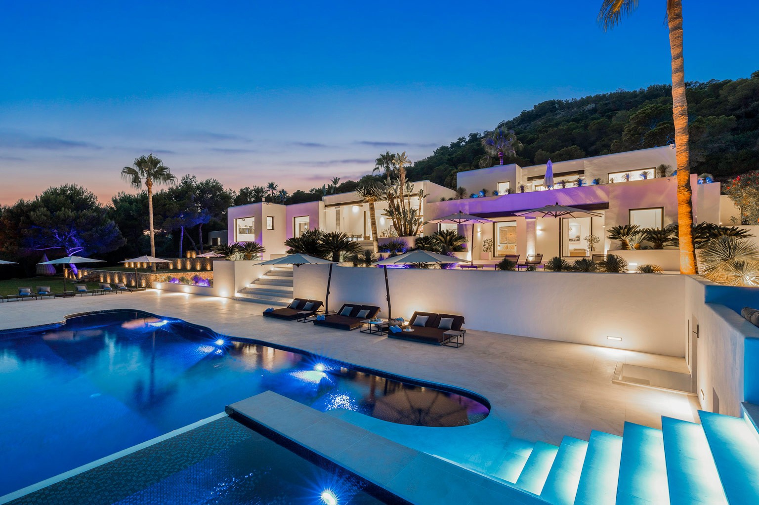 Exclusive luxury villa in Ibiza with a glowing pool at night, surrounded by lounge seating and palm silhouettes—a private VIP estate property holiday retreat for weddings and events, offering privacy.