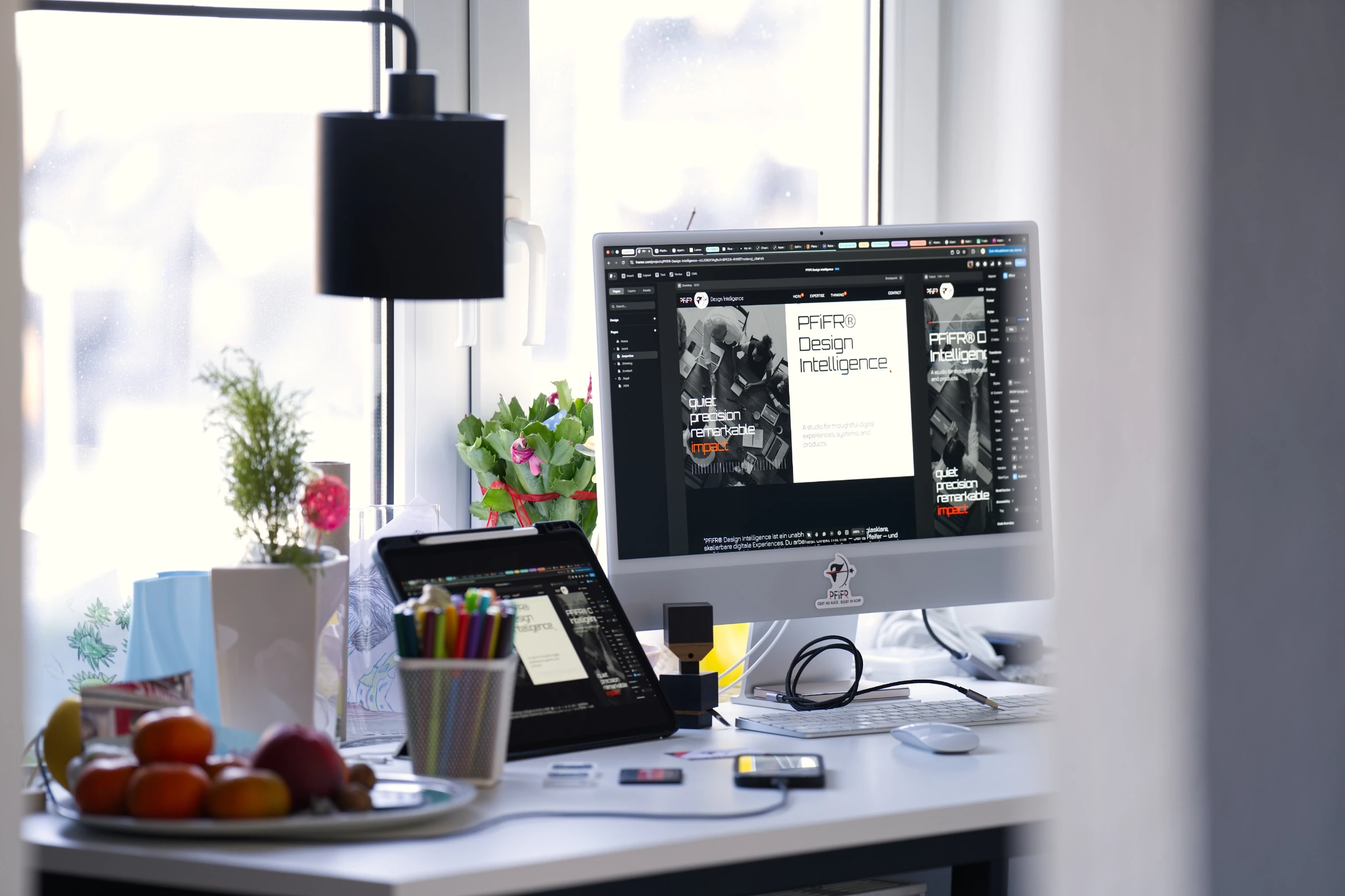 Seitliche Perspektive eines kreativen Arbeitsplatzes mit großem Monitor, Tablet und Tastatur, auf dem die Benutzeroberfläche von „PFiFR Design Intelligence“ zu sehen ist, ergänzt durch Pflanzen, Stifte und persönliches Arbeitsmaterial.
