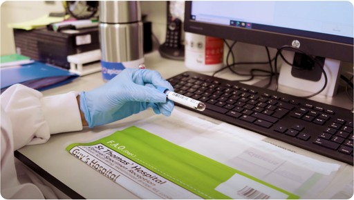 A person wearing blue medical gloves holds a test tube near a computer keyboard on a laboratory desk, with hospital documents and laboratory equipment in the background.
