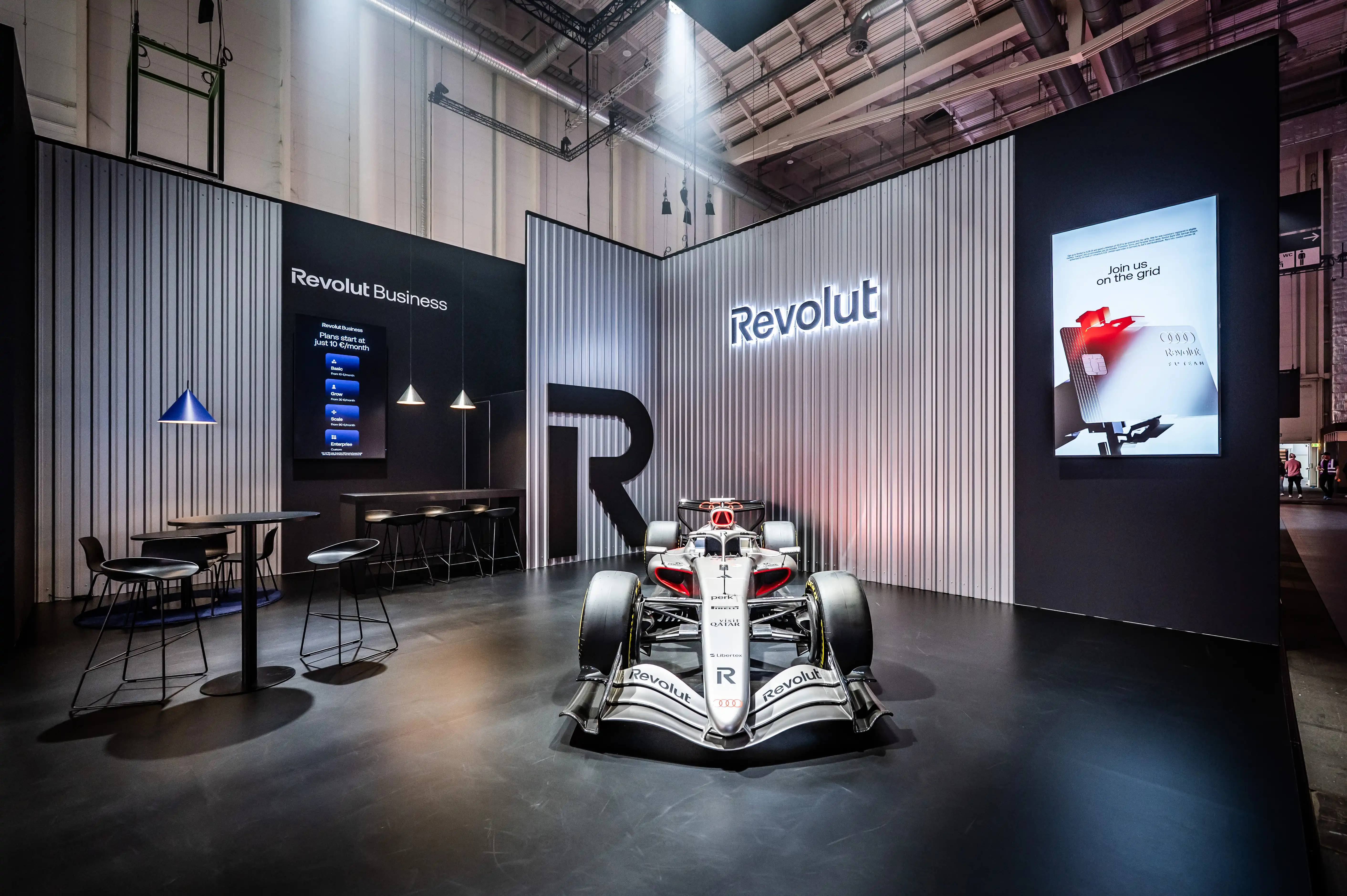 Revolut booth with Formula 1 car, seating area, screens and metallic striped walls in the exhibition hall.