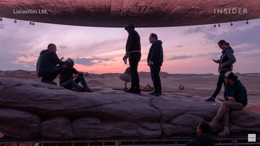 A group of people gathered under a rocky overhang during sunset, with a colorful sky in the background.