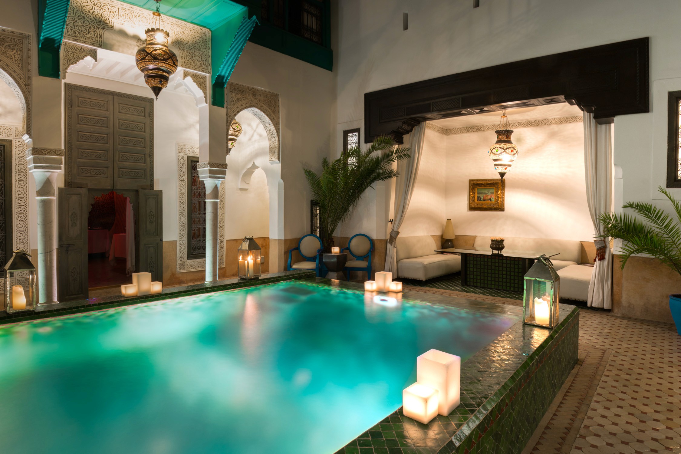 A luxury spa courtyard features a turquoise pool with glowing lanterns and two lounge chairs under a draped canopy.