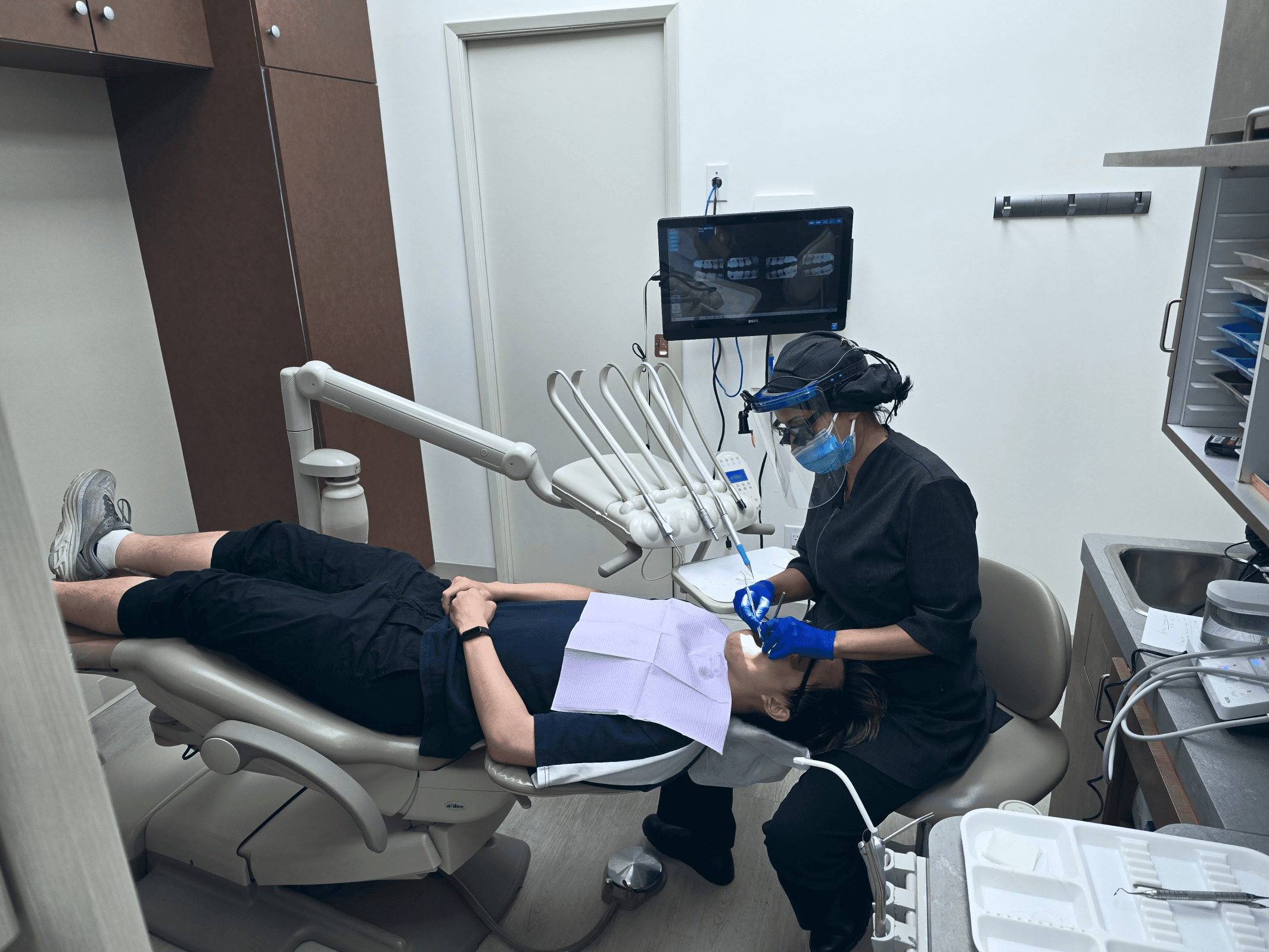 A dental professional wearing protective gear and a mask performing a dental procedure on a patient lying in a dental chair.