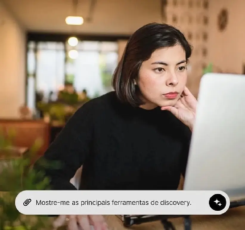 Uma mulher sentada em uma mesa, concentrada em seu laptop, com plantas ao fundo e um interior casual e moderno.