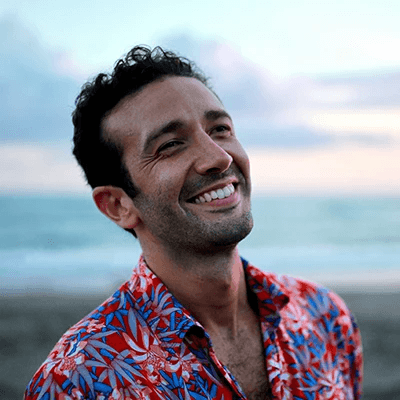 Man smiling while wearing a colorful floral shirt, standing on a beach at sunset. The background shows a calm ocean and a soft, pastel sky.