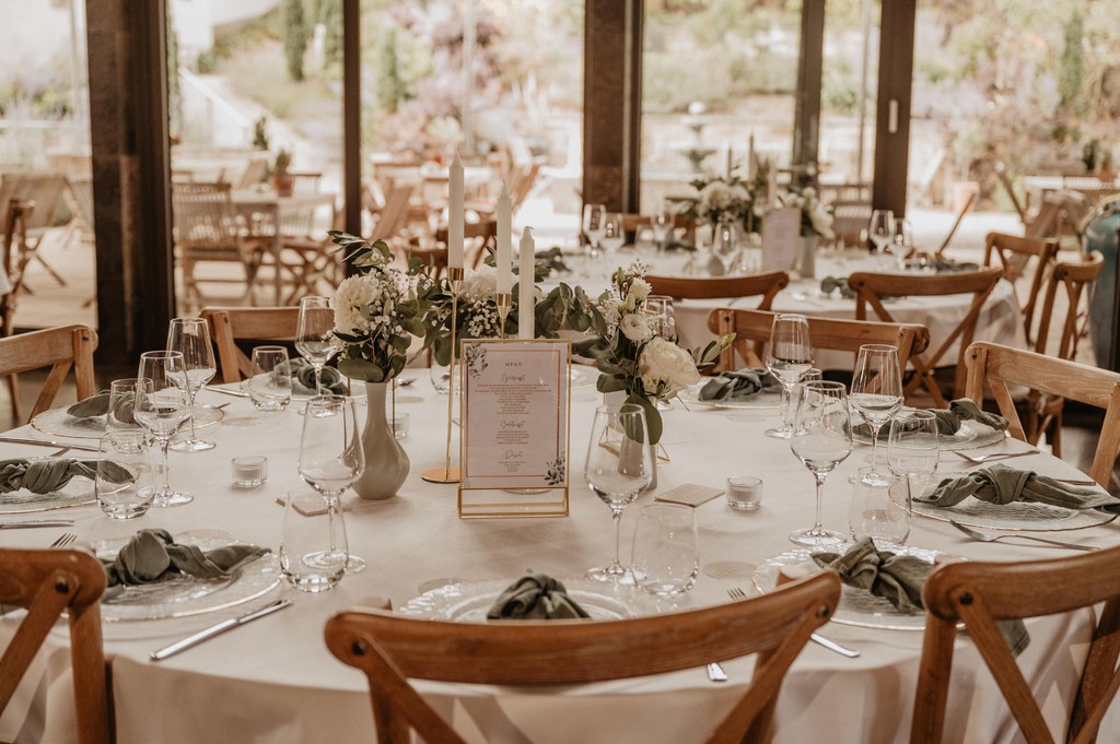 Festlich gedeckter runder Hochzeitstisch mit weißen Blumen, Eukalyptus, Menükarten in Goldrahmen und Kerzenständern
