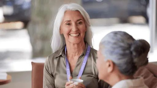 A smiling older woman with white hair wearing a lanyard, engaged in conversation with another person in a professional setting