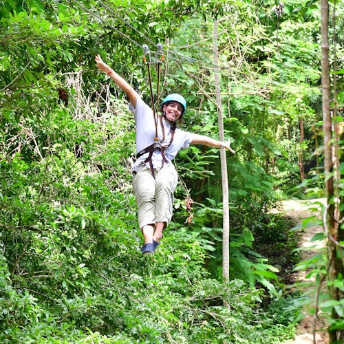 一个人微笑着通过郁郁葱葱的绿色森林进行高空滑索，身穿安全带和头盔。