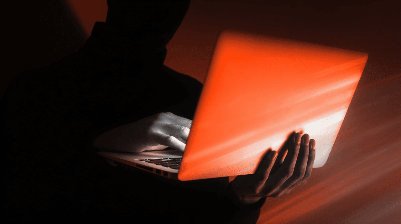 Silhouette of a person using a laptop with a bright orange glow from the screen.