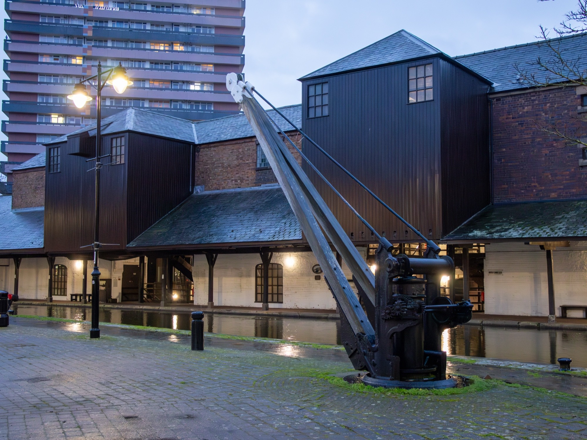 A canal-side crane beside warehouse buildings at the basin, lamps lit at dusk.