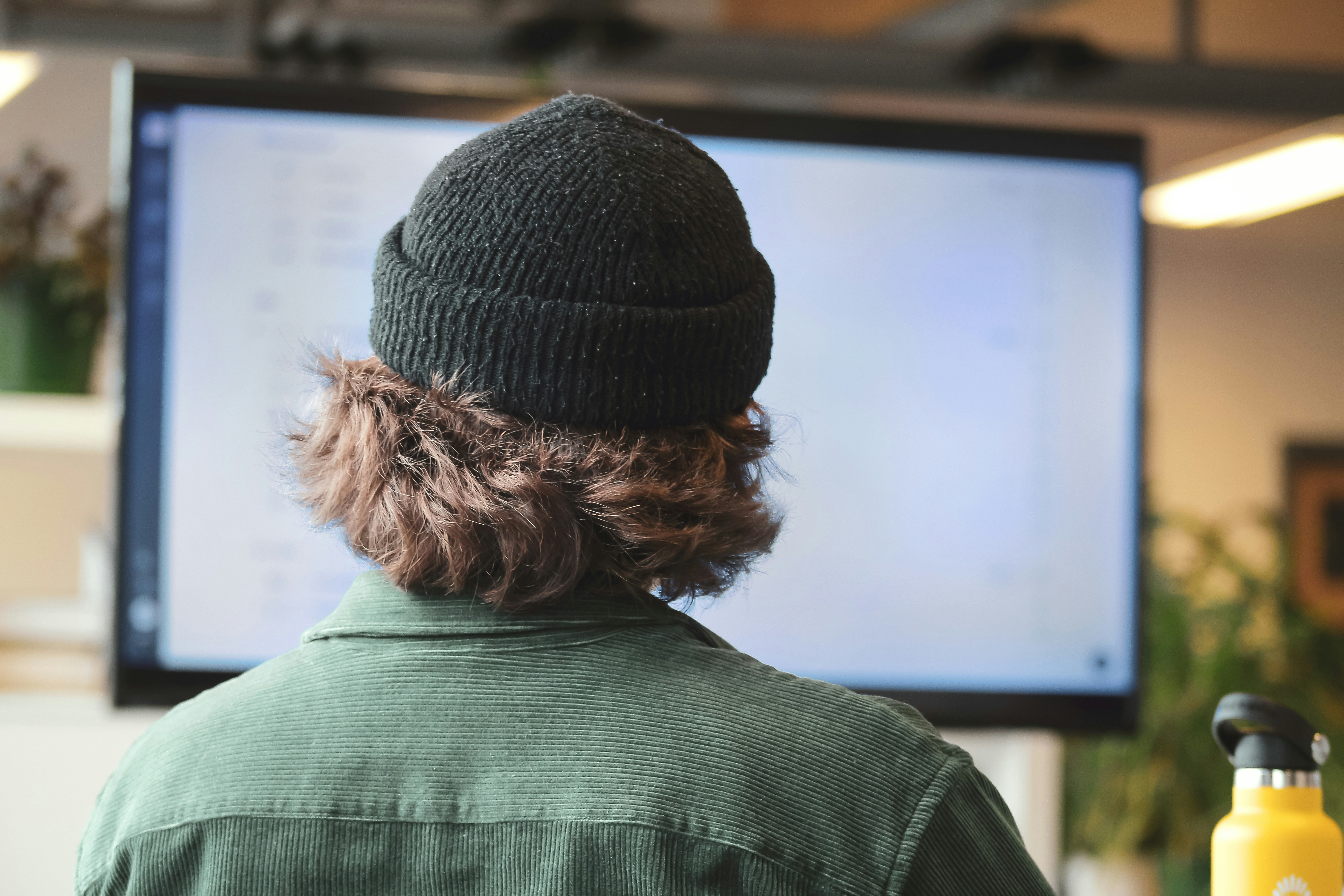 Backside of a person with a had and curly hairs sits in front of a screen in a room