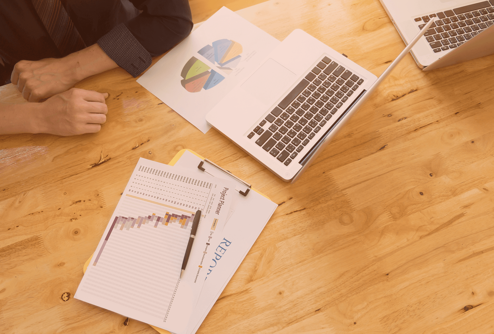 Overhead view of a marketing professional working at a wooden desk with a laptop, a colorful pie chart, and a printed project planner report.
