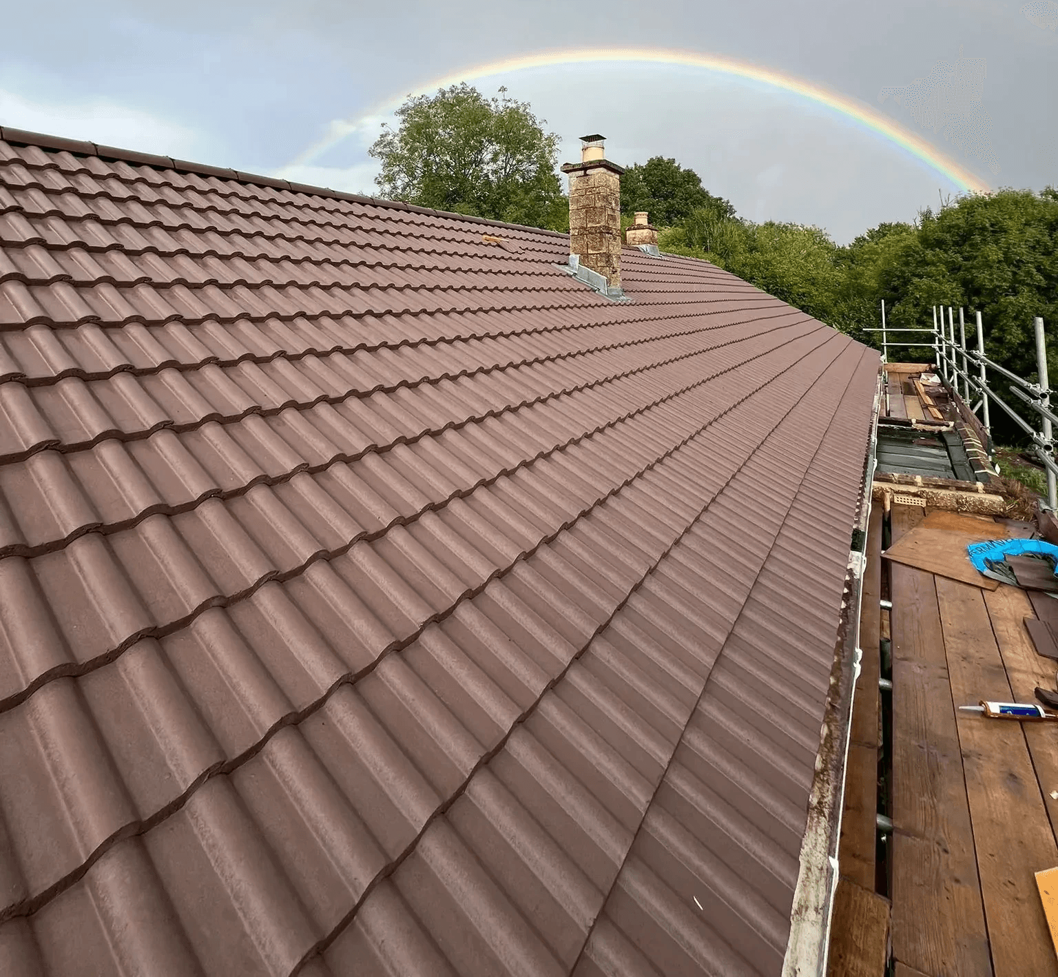 Close-up of a brown tiled roof with chimney under a vibrant rainbow in the sky.