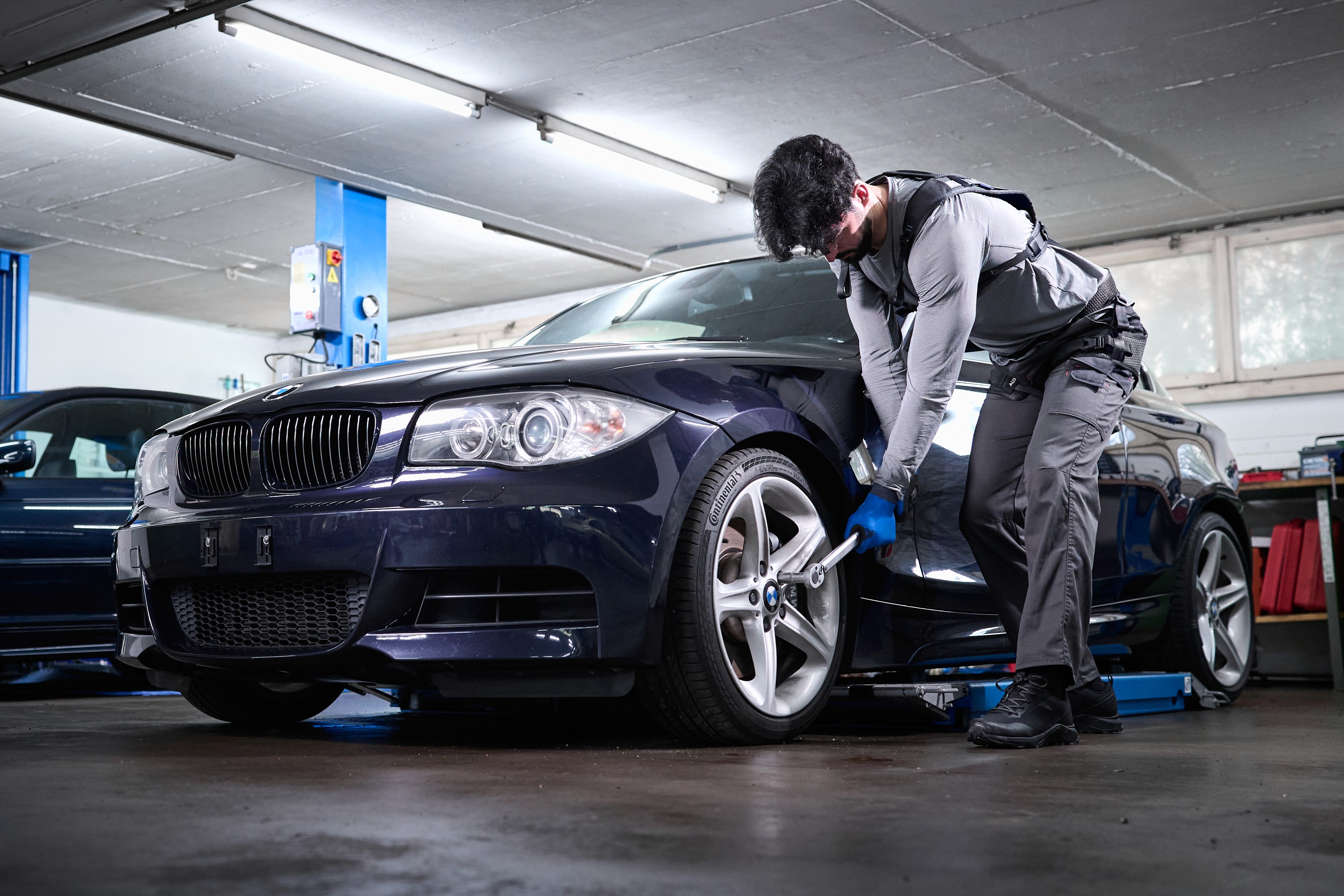 Auto mechanic wearing an Auxivo Liftsuit exoskeleton in a garage, tightening a wheel nut on a BMW car to illustrate the prevention of back pain at work.
