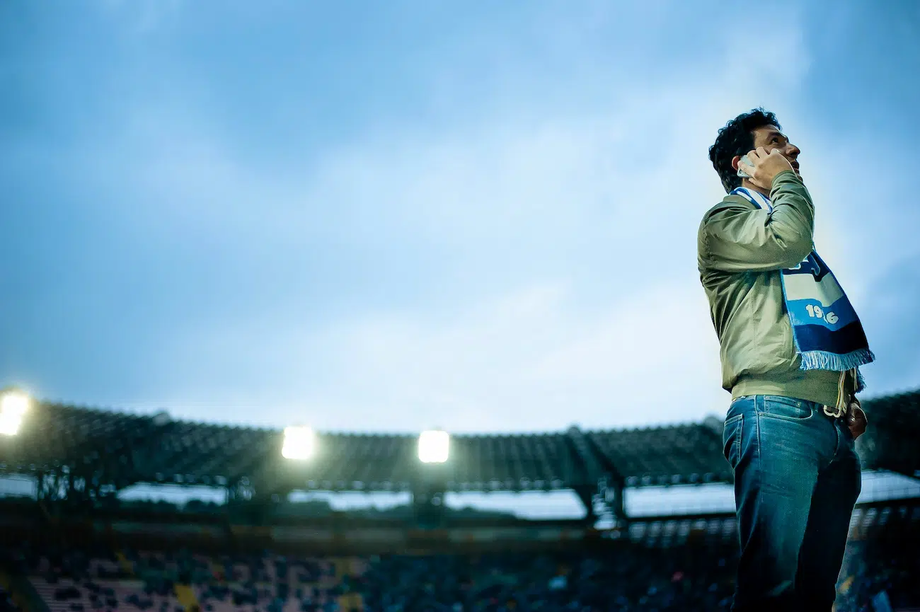 A man in a stadium wearing a sports scarf and holding a phone to his ear, standing against a backdrop of bright stadium floodlights and a partially filled seating area under an open sky. The image captures the essence of modern sports events and communication, aligning with the concept of digital tools for league and tournament organisation, tailored for clubs and event organisers.