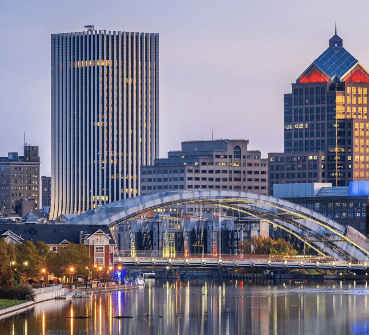 Downtown Rochester, New York skyline at dusk featuring the Genesee River, illuminated city lights, and the modern arched bridge over the river.