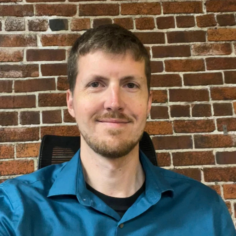 Uma pessoa com cabelo curto sorri suavemente, vestindo uma camisa de botões verde-azulada, sentada em um ambiente interno diante de uma parede rústica de tijolos vermelhos.