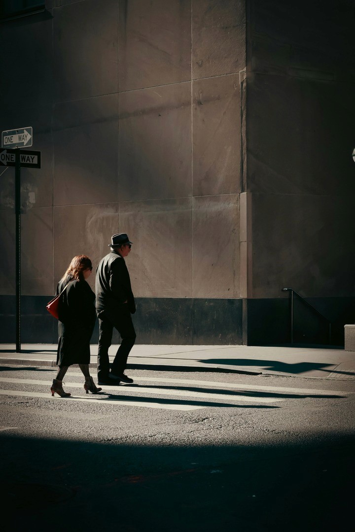 inematic street photography of two figures silhouetted as they step through a bright patch of sunlight on an otherwise shadowed city street corner.