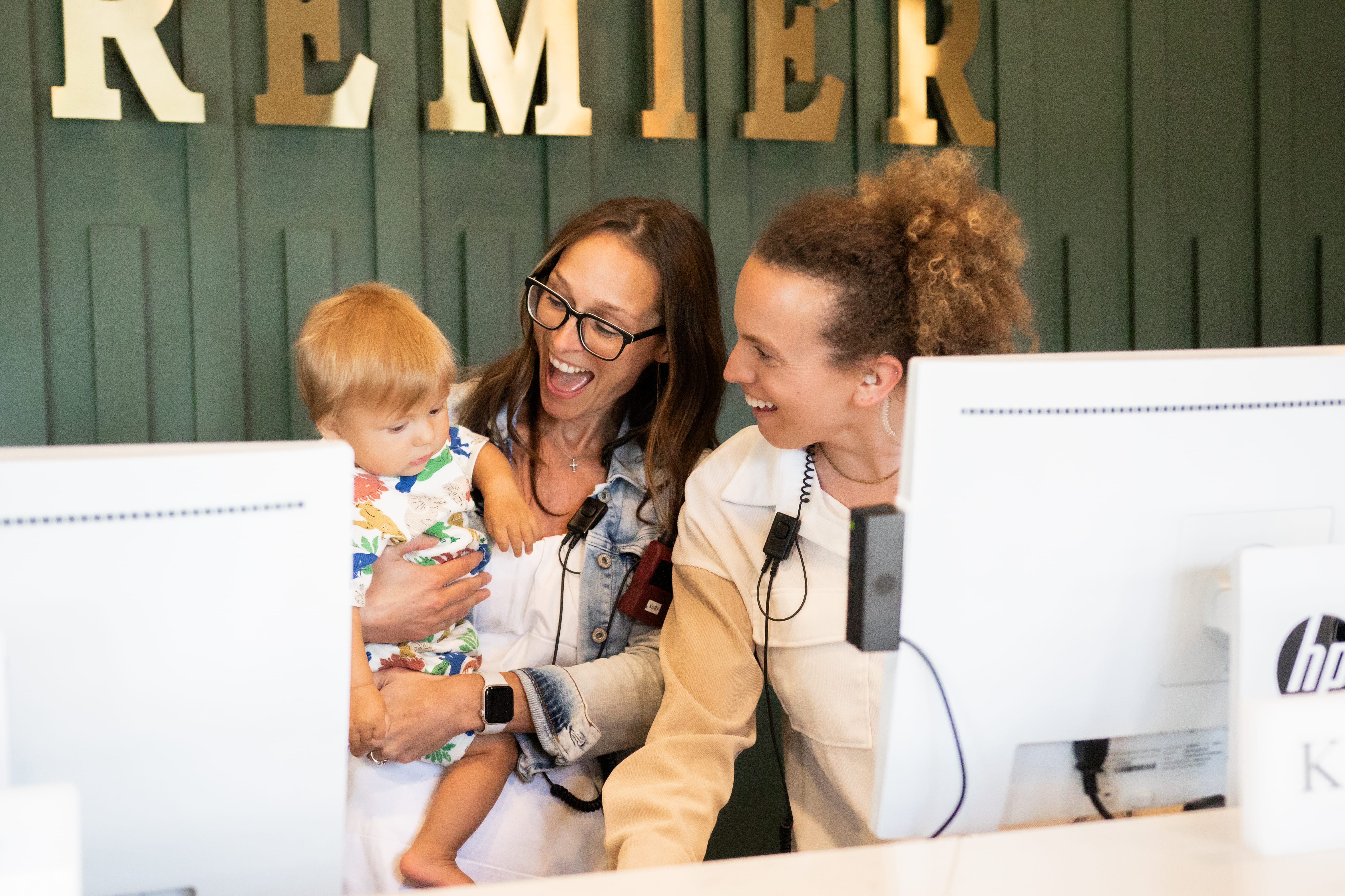 Two smiling employees holding a child and sitting at the front desk of the Premier office.