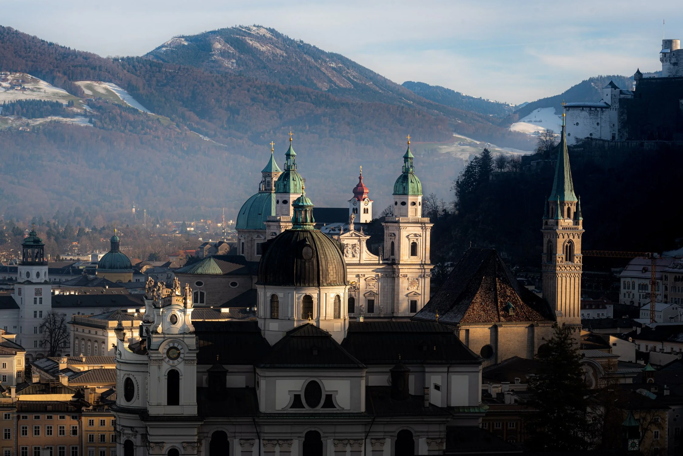 A panoramic view of a historic city with domed and bell towers in front of snow-dusted mountains under a blue sky.