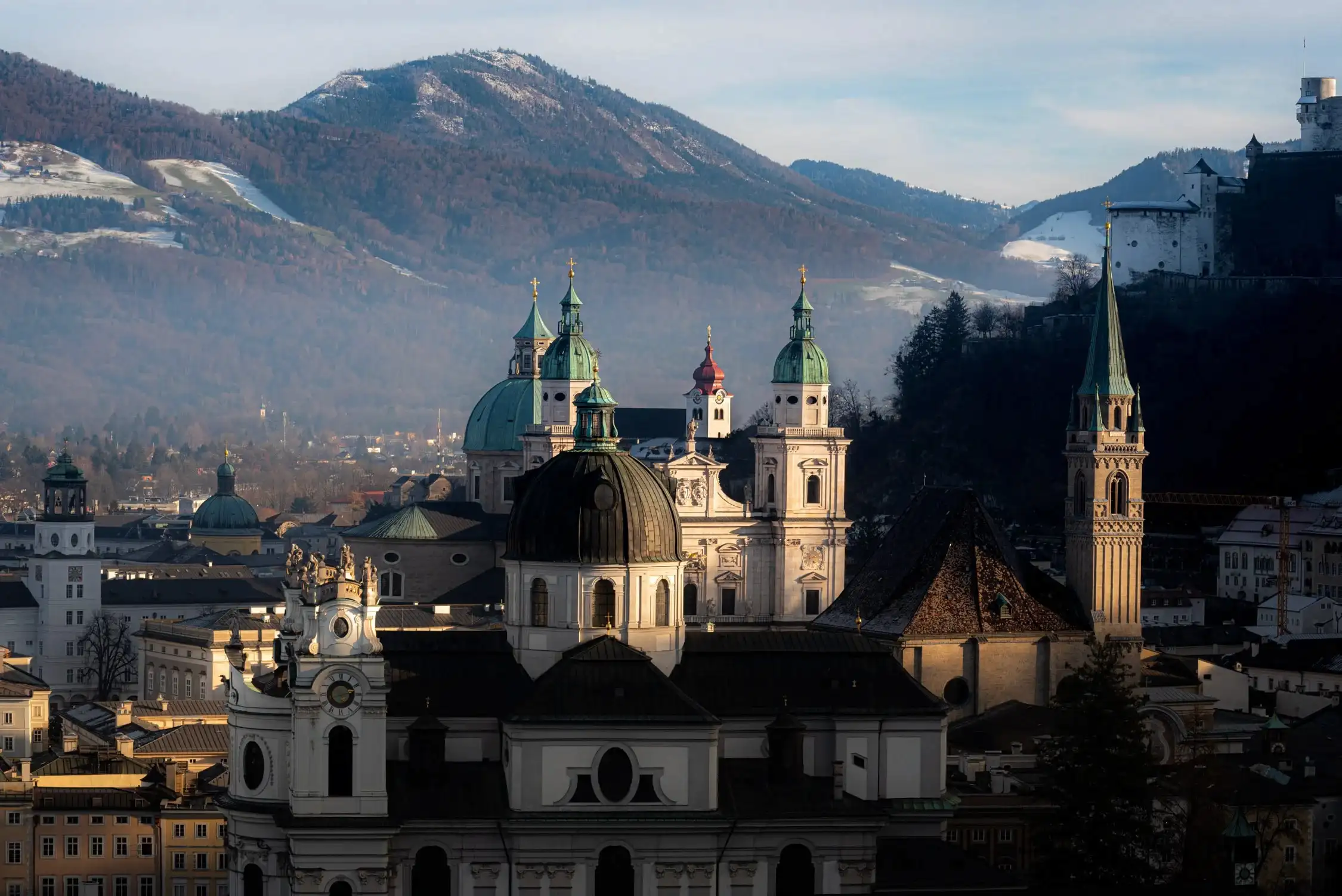 A panoramic view of a historic city with domed and bell towers in front of snow-dusted mountains under a blue sky.