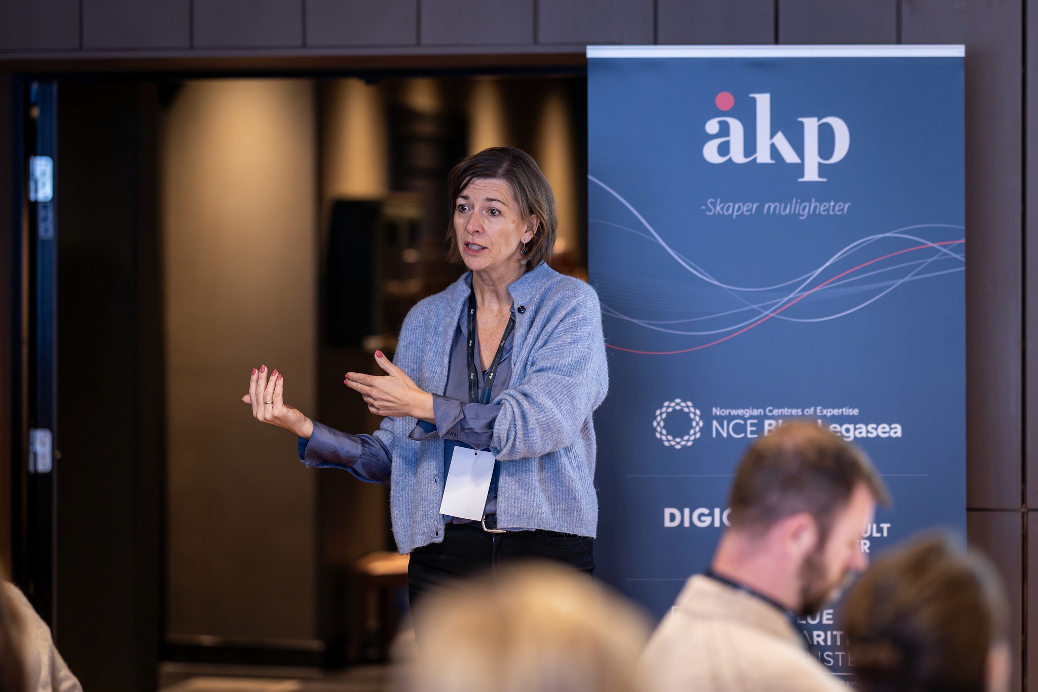 A woman with blonde hair smiles in a social setting, with people blurred in the background. Bright window visible.
