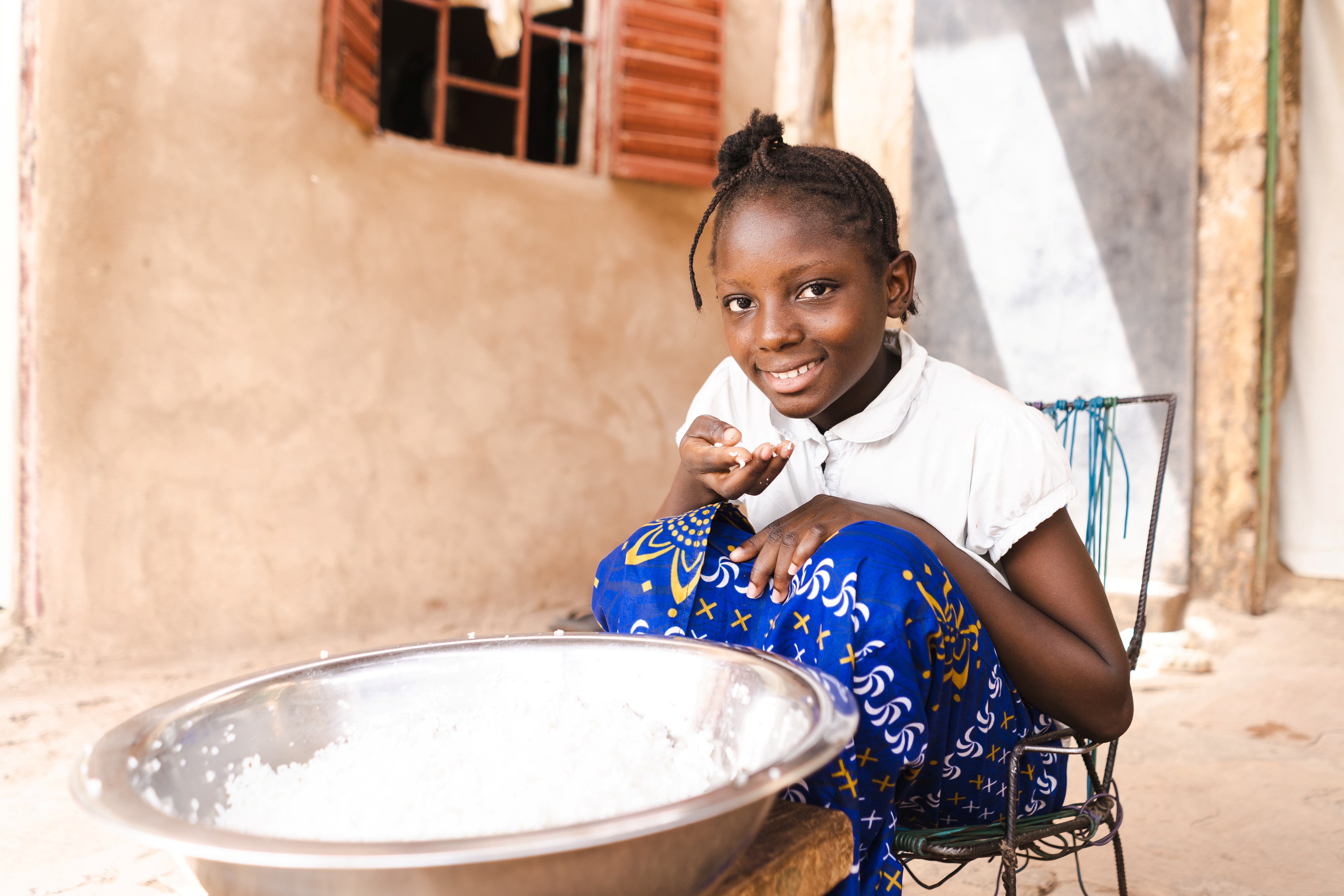 A young girl in a white shirt and blue patterned skirt smiles while holding a handful of food next to a large metal bowl, sitting outside her home in a developing country.